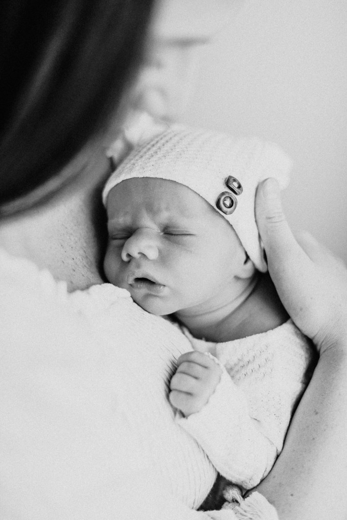 A black and white image of a mother holding a sleeping newborn baby, who is wearing a knitted hat and gently touching its hand against the mother's chest.